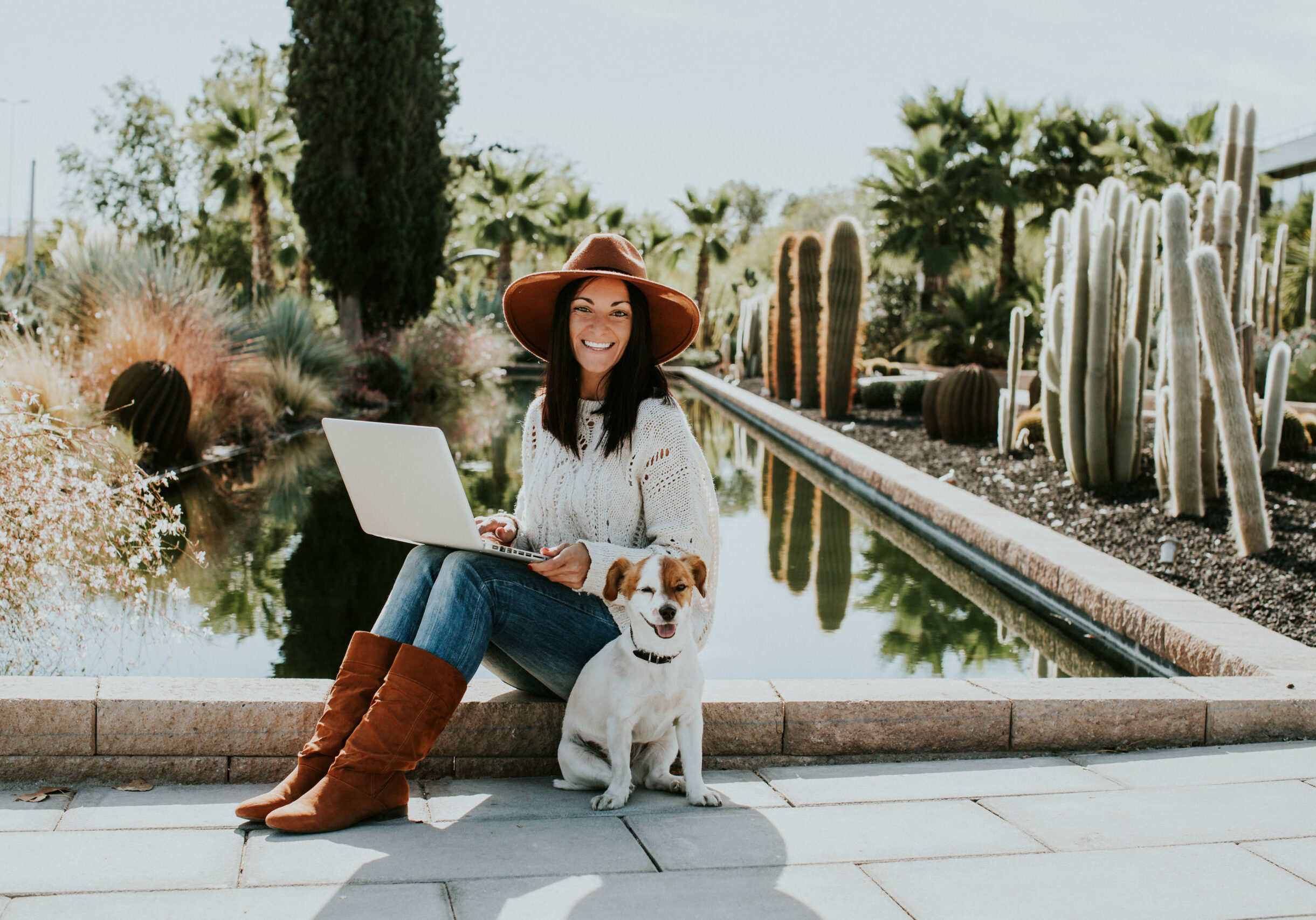 Pretty young woman working outdoors with her adorable dog jack rusell in a park surrounded by cactus and a small pond.